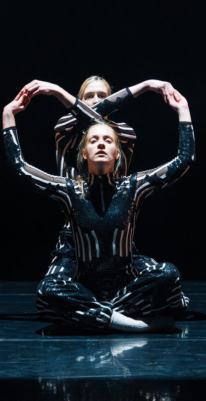 Two figures in striped body suits on a black background, sit on the floor facing forwards. Their arms make two circles