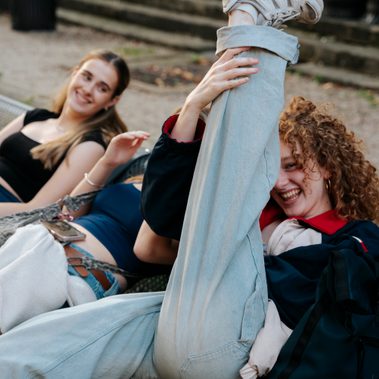 Three young people are sat on a bench at an angle to the camera, one of them with red hair has raised her leg high into the air. They are all laughing and smiling