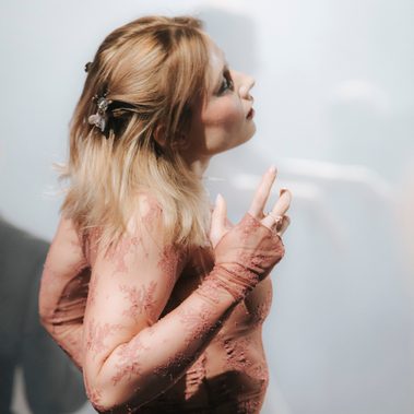 A person with long blond hair and lacey top stands sideways to the camera, looking slightly upwards