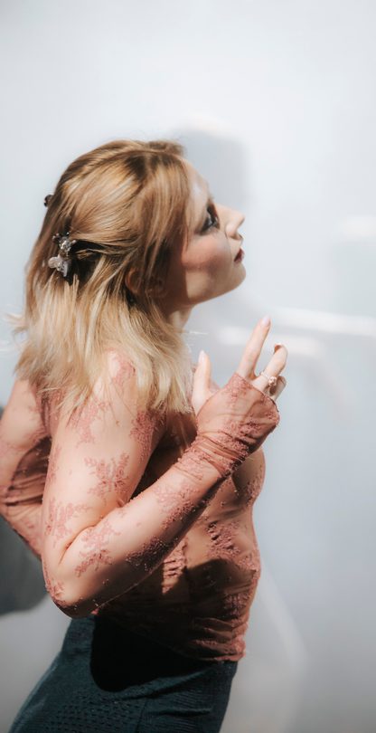 A person with long blond hair and lacey top stands sideways to the camera, looking slightly upwards