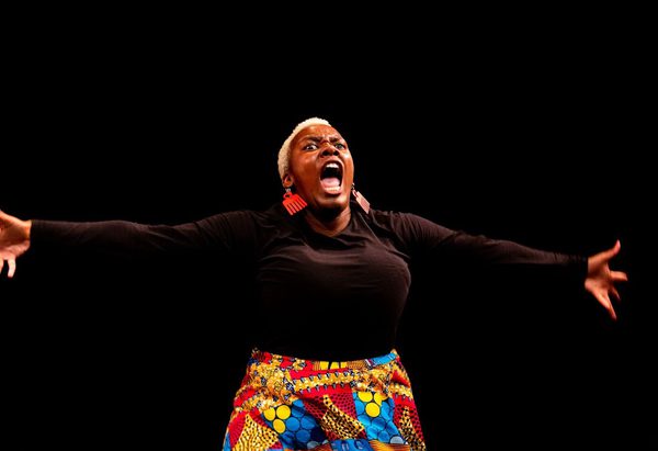 Against a black background, a person with short, curly white hair, wearing large red earrings, a black long-sleeve top, and a colourful patterned skirt, are shown from the hips up, stretching their arms to the sides and opening their mouth in a scream or alarmed expression.