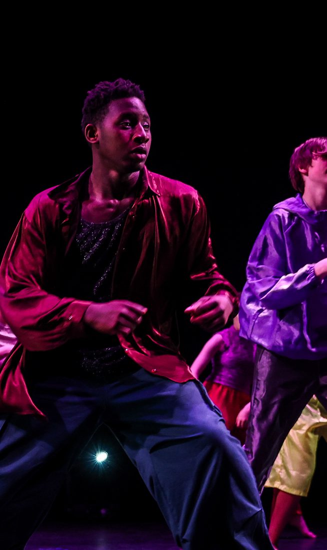 A group of young dancers between 14 and 19 on a stage against a black background. They are wearing satin clothing in a mixture of rich colours.