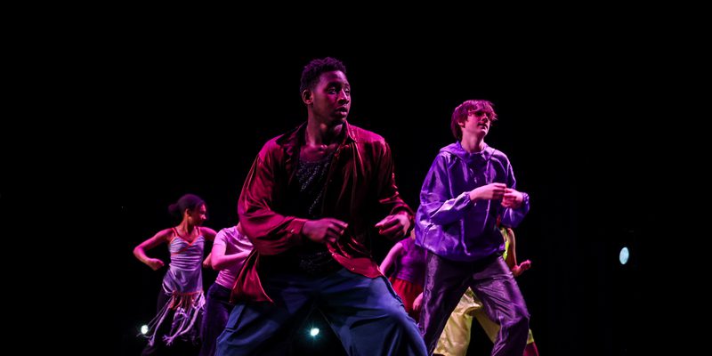 A group of young dancers between 14 and 19 on a stage against a black background. They are wearing satin clothing in a mixture of rich colours.