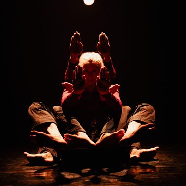 Two dancers sit one behind the other on a dark stage, illuminated by a warm overhead light. Their bodies overlap to create a multi-limbed silhouette, with several pairs of hands raised in flat, palm-forward gestures and their bare feet fanned out at the base.