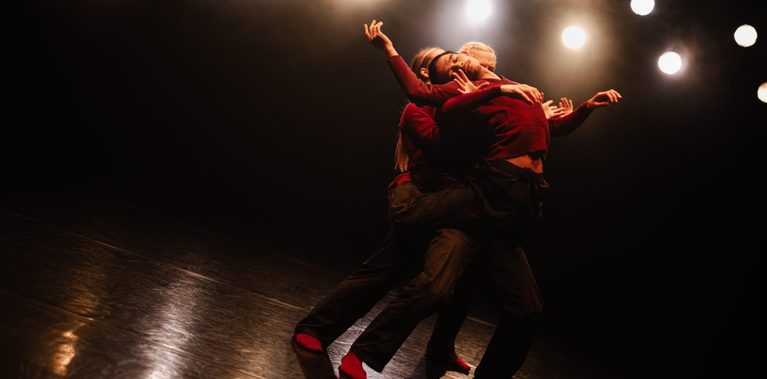 Three dancers wearing dark red tops and black trousers are grouped closely together on a dark stage, leaning back at a sharp angle. Their arms are intertwined and reaching outward in fluid, expressive motions. The scene is illuminated by a series of warm, out-of-focus stage lights in the background, which reflect off the high-shine floor.