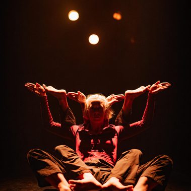 Two dancers sit back-to-back in a seated position on a dark stage, illuminated from above by several warm, glowing lights. Their legs are spread wide toward the foreground, and their multiple arms are raised and fanned out symmetrically, with hands held flat and palms facing upward, creating an intricate silhouette against the darkness.