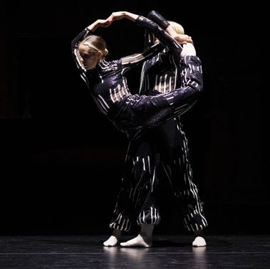 Two dancers in shimmering, black-and-white striped costumes perform a dynamic lift on a dark stage. The dancer in the foreground is lifted horizontally with their legs tucked and back arched. Their arms reach upward to meet the arms of the supporting dancer, forming a circular, frame-like shape with their hands against the black background.