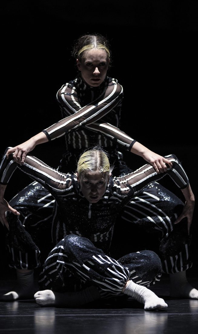Two dancers wearing shimmering black-and-white striped costumes perform on a dark stage under a spotlight. One dancer is seated cross-legged on the floor, while the second stands closely behind them, leaning forward so their arms cross over the seated dancer's shoulders to create a symmetrical, geometric shape.