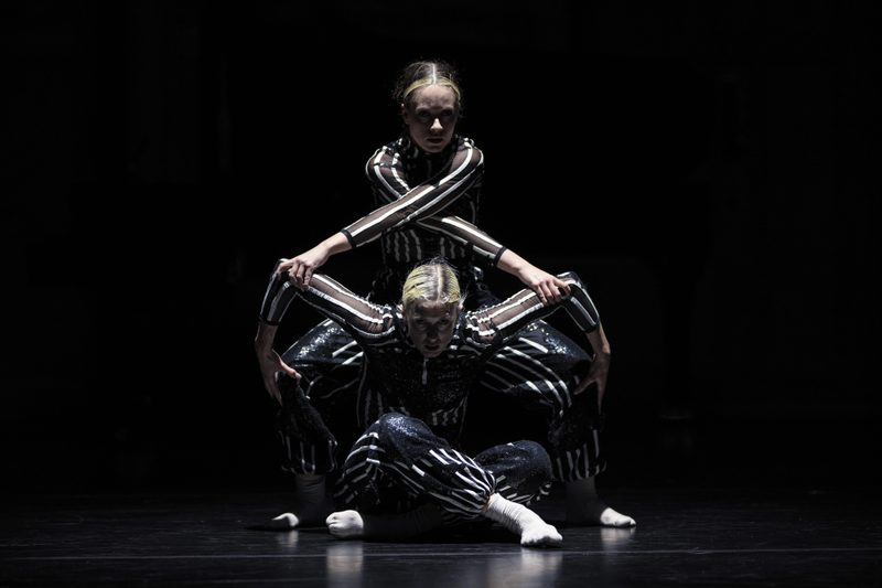 Two dancers wearing shimmering black-and-white striped costumes perform on a dark stage under a spotlight. One dancer is seated cross-legged on the floor, while the second stands closely behind them, leaning forward so their arms cross over the seated dancer's shoulders to create a symmetrical, geometric shape.