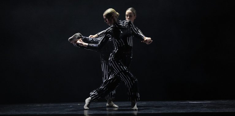 Gemini said Splice Production (Tangled Silhouette) Two dancers in shimmering black-and-white striped costumes perform a complex entanglement on a dark stage. One dancer leans forward with their leg extended backward, while the other stands closely behind, their bodies overlapping to create a multi-limbed, layered silhouette under dramatic stage lighting.