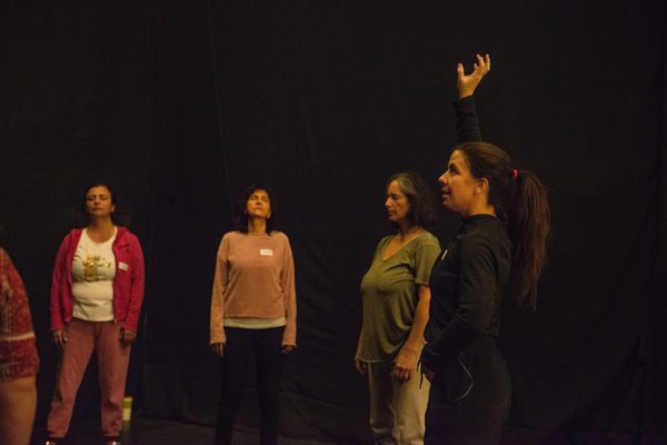 Four people stand spaced apart against a dark backdrop, appearing to take part in a movement exercise. In the foreground, a woman wearing black clothing raises one arm overhead while the others stand upright, with their eyes close.