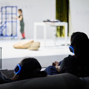 A photo of the backs of audience members, with headphones on, watching child performers on stage.
