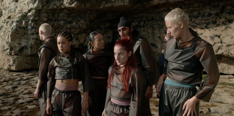 A group of six performers stands close together against a backdrop of rock cliffs. They wear matching dark, translucent, oversized tracksuits over various base layers. The group is positioned in a staggered formation, looking in different directions with serious, focused expressions. The natural lighting is bright but soft, highlighting the textures of the stone and the thin material of their costumes.