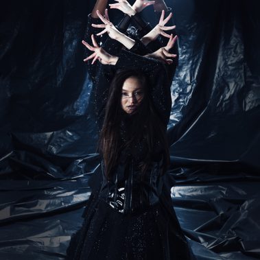 A performer stands in front of a textured black backdrop. Above her head her hands are crossed and with her fingers spread, above her arms another two sets of hands, the owners unseen, pose in a similar way creating a crown like image. Stacked at the very top, one last pair of hands makes a circular symbol like shape.