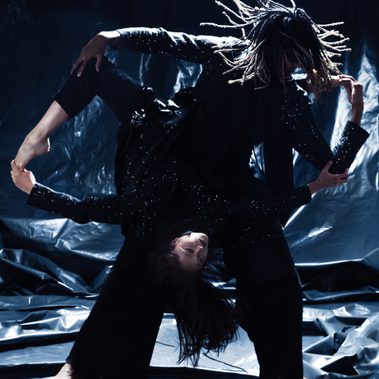 Two dancers are held in an intricate lift. One stood while the other has there legs wrapped around them so they can hold themselves upside down. Their arms and the suspended dancers legs make a circle like shape as their hair flies around them.