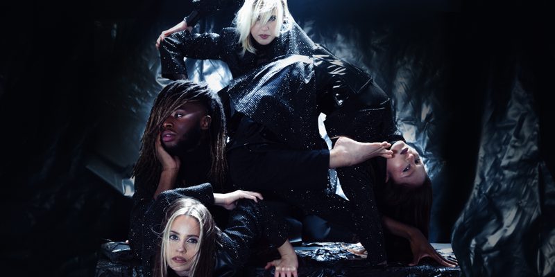Four performers pose against a textured black backdrop. One dancer is in a bridger pose with their left foot resting against her shoulder. The other three are stacked to her left in different striking poses. They are all wearing black sparkling costumes.