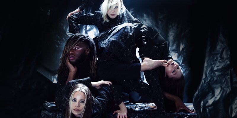 Four performers pose against a textured black backdrop. One dancer is in a bridger pose with their left foot resting against her shoulder. The other three are stacked to her left in different striking poses. They are all wearing black sparkling costumes.