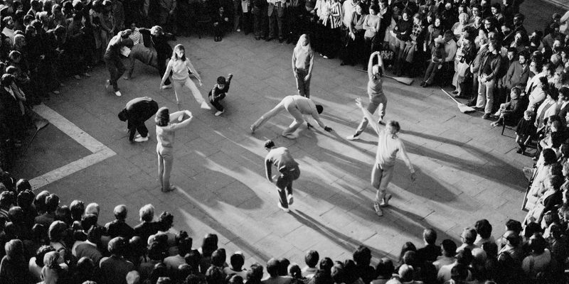 Black and white image of Merce Cunningham's Event performed in Venice's St Mark's Square in 1972. The image is taken from a bird's eye view. 7 dancers wearing light colour costumes and 5 wearing darker costumes and encircled by a large audience.