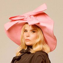 Image of Paloma Faith Blomfield. Paloma is a white woman with blonde hair and wears a large pink hat with a big bow on the front. Paloma wears s brown jacket.