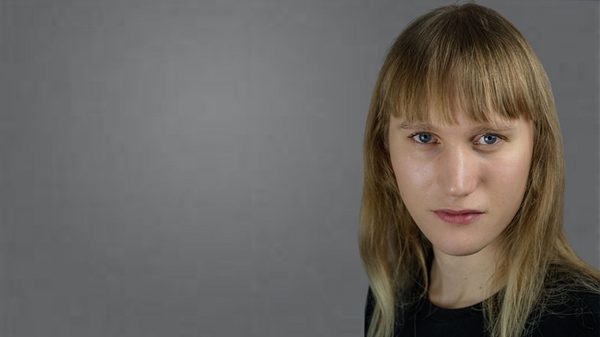 A white blond person is looking in the camera in front of a grey background. They have long blond hair and a fringe and are wearing a black t-shirt.