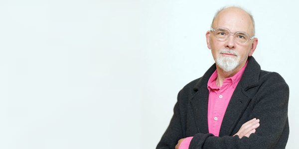 Image of Bob Lockyer. Bob looks into the camera with his arms folded. He is wearing a pink shirt with a black cardigan over the top.