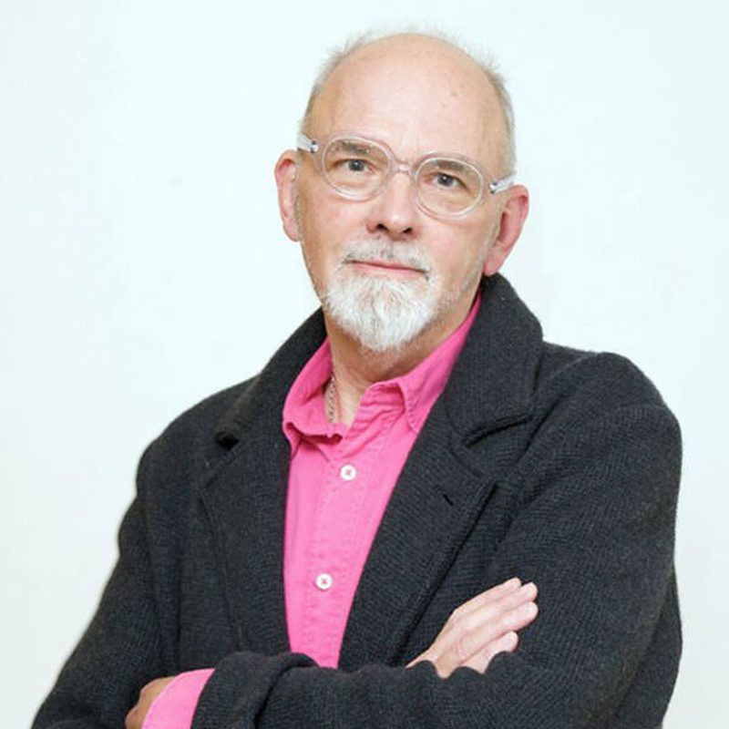 Image of Bob Lockyer. Bob looks into the camera with his arms folded. He is wearing a pink shirt with a black cardigan over the top.