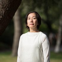 a lady is standing in a garden wearing a white top, looking just above at the camera. She has short brown hair.