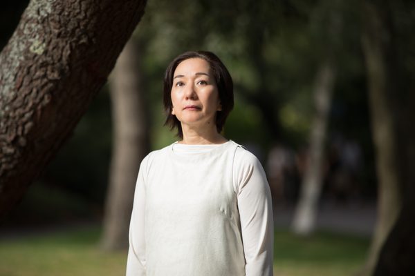 a lady is standing in a garden wearing a white top, looking just above at the camera. She has short brown hair.