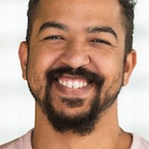 A headshot of a young man with a big smile and a goaty beard and short brown hair. He is wearing a pink t-shirt and is in front of a blurred window.