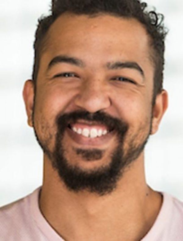 A headshot of a young man with a big smile and a goaty beard and short brown hair. He is wearing a pink t-shirt and is in front of a blurred window.