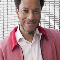 Image of Kenneth Olumuyiwa Tharp. Kenneth is a black man and wears a dark pink jacket with a lighter pink shirt underneath.