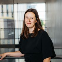 A white woman is looking at the camera smiling. She has brown hair down to under her shoulders and is wearing a dark blue dress.