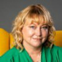 A blond woman with mid-length hair is looking in the camera and smiling. She is wearing a green top and is sat on a yellow chair.