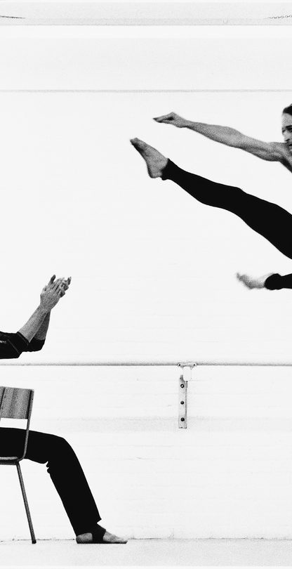 Black and White photo of Robert (Bob) Cohan in rehearsal with a member of the London Contemporary Dance Theatre. Cohan is on a chair clapping at a performer in the air with black leggings and kicking his leg forward