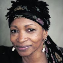 Portrait of Bonnie Greer. She is a black woman wearing a head scarf with some hair showing and has white / silver earrings.
