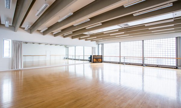 Image of an empty studio with widows on the right hand side and a ballet barre across the windows and mirrors on the left wall. There is a piano in the corner