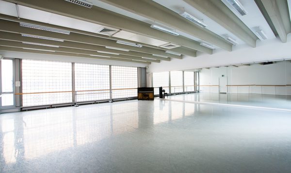 Image of an empty studio with mirrors on the right hand side and widows on the left with ballet barres across the window and a piano in the corner