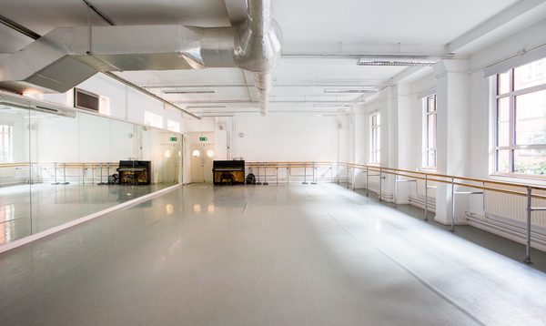Image of an empty studio with windows on the right hand side with ballet barres across the wall, a piano on the far wall and mirrors on the left hand side