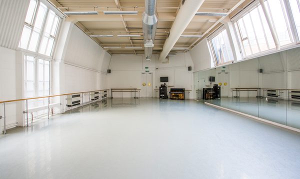 Image of an empty studio with windows and ballet barres on the left hand side, a piano by the far wall and mirrors on the right hand side