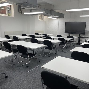 a lecture room with white desks facing a back wall. There is a TV screen on a stand on wheels at the end of the room