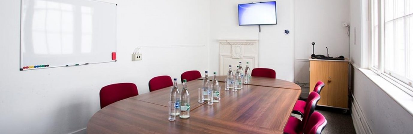 A photo of our meeting room, there is a long wooden table with round ends in the middle of a white-painted room. There are 8 red chairs around the table and some water bottles and glasses on the table. There is a screen on the far right hand side wall and a whiteboard on another wall.