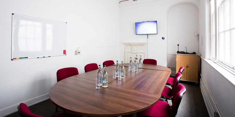 A photo of our meeting room, there is a long wooden table with round ends in the middle of a white-painted room. There are 8 red chairs around the table and some water bottles and glasses on the table. There is a screen on the far right hand side wall and a whiteboard on another wall.