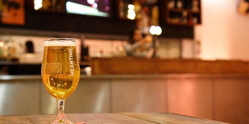 A photo of the theatre bar at The Place. We see a wooden table with a pint of meantime beer on it. The bar is in the background.