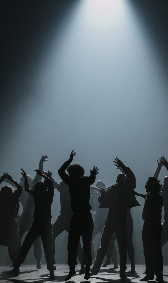 Silhouettes of a group of dancers performing on a dark stage beneath a single bright spotlight. Their arms and bodies are extended in expressive poses, creating a dramatic, high-contrast scene against the illuminated background.