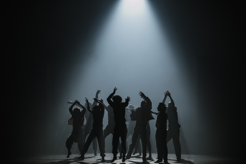 Silhouettes of a group of dancers performing on a dark stage beneath a single bright spotlight. Their arms and bodies are extended in expressive poses, creating a dramatic, high-contrast scene against the illuminated background.