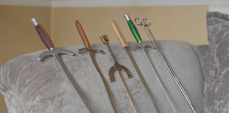Six swords of different types lean against the armrest of a grey velvet sofa. At their base lies a metal swallowing chain with two ball-shaped ends. The scene is quiet and still, with a calm, almost unsettling atmosphere that contrasts with the dangerous nature of the objects.