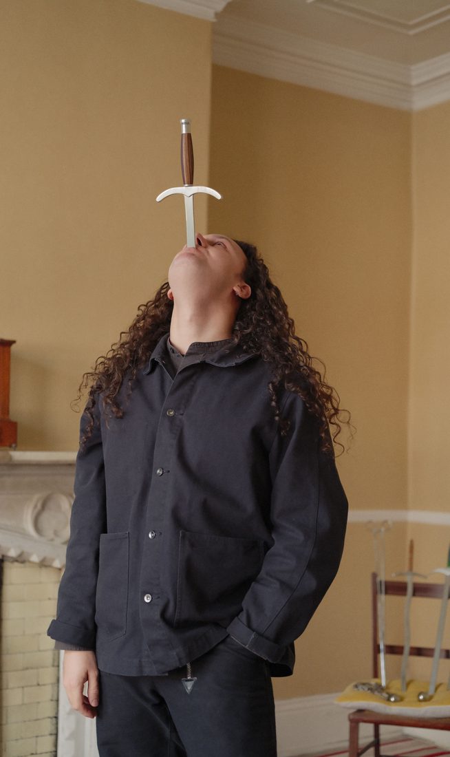 Photo of Tom Cassani in the middle of swallowing a sword, with the handle and upper section of the sword still visible. He is looking upward, has long curly brown hair, and is wearing a black long-sleeved button-up shirt. The image is taken indoors against plain yellow walls.