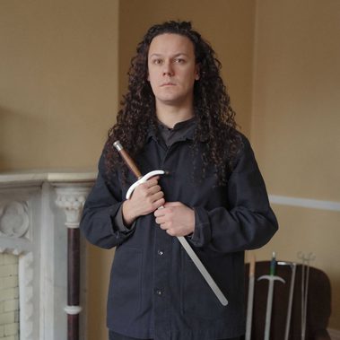 Portrait of Tom Cassani shown from the waist up, standing and facing the camera. He holds a sword with both hands across the front of his body. He has long, curly dark hair and is wearing a black long-sleeved button-up shirt. The photograph is taken indoors against plain yellow walls, with part of a fireplace visible behind him and four different swords displayed in the background.