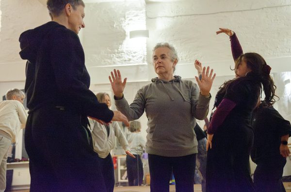Three people participate in a movement workshop in a bright studio with white walls and mirrors. At the centre, a woman with grey hair in a grey hoodie holds both hands raised at chest height while two other people face them, each striking expressive, dance-like poses.