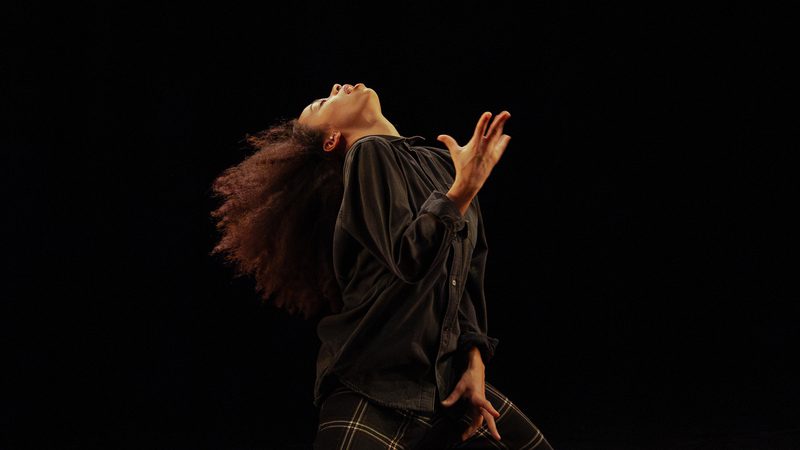 Against a black background, a person with long, curly brown hair wearing a black shirt and black checkered trousers leans their head back, one arm bent at the elbow and the other resting along their side, both hands with fingers slightly spread, and knees bent.